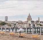 A picture of a beekeeper tending to one beehive (out of four) on a roof of a building with the skyline of a city (Paris) on the background. Our experimental apiary on the roof of Sorbonne university, the Jussieu campus.